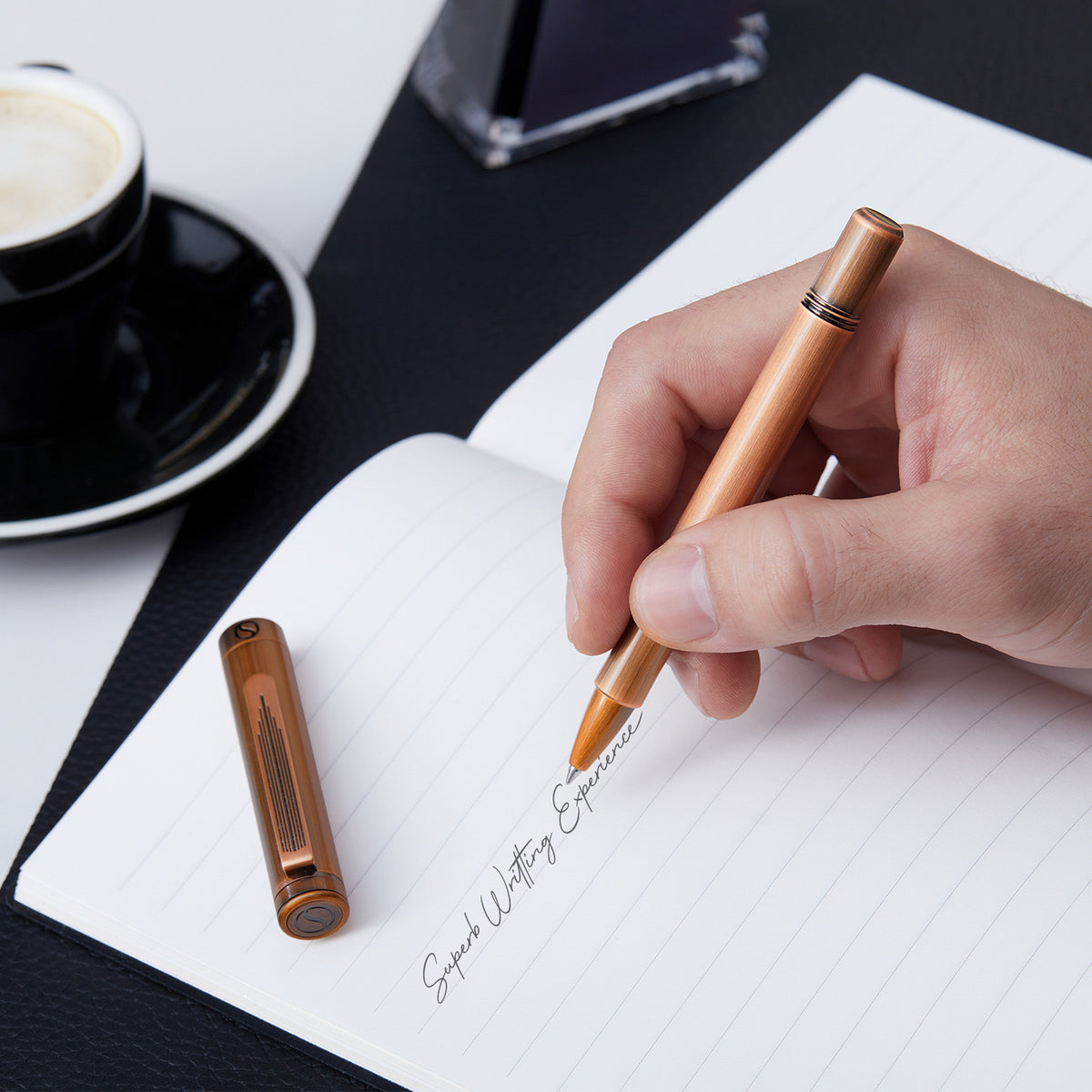 Hand holding a copper pen over a notebook with a cup of coffee in the background