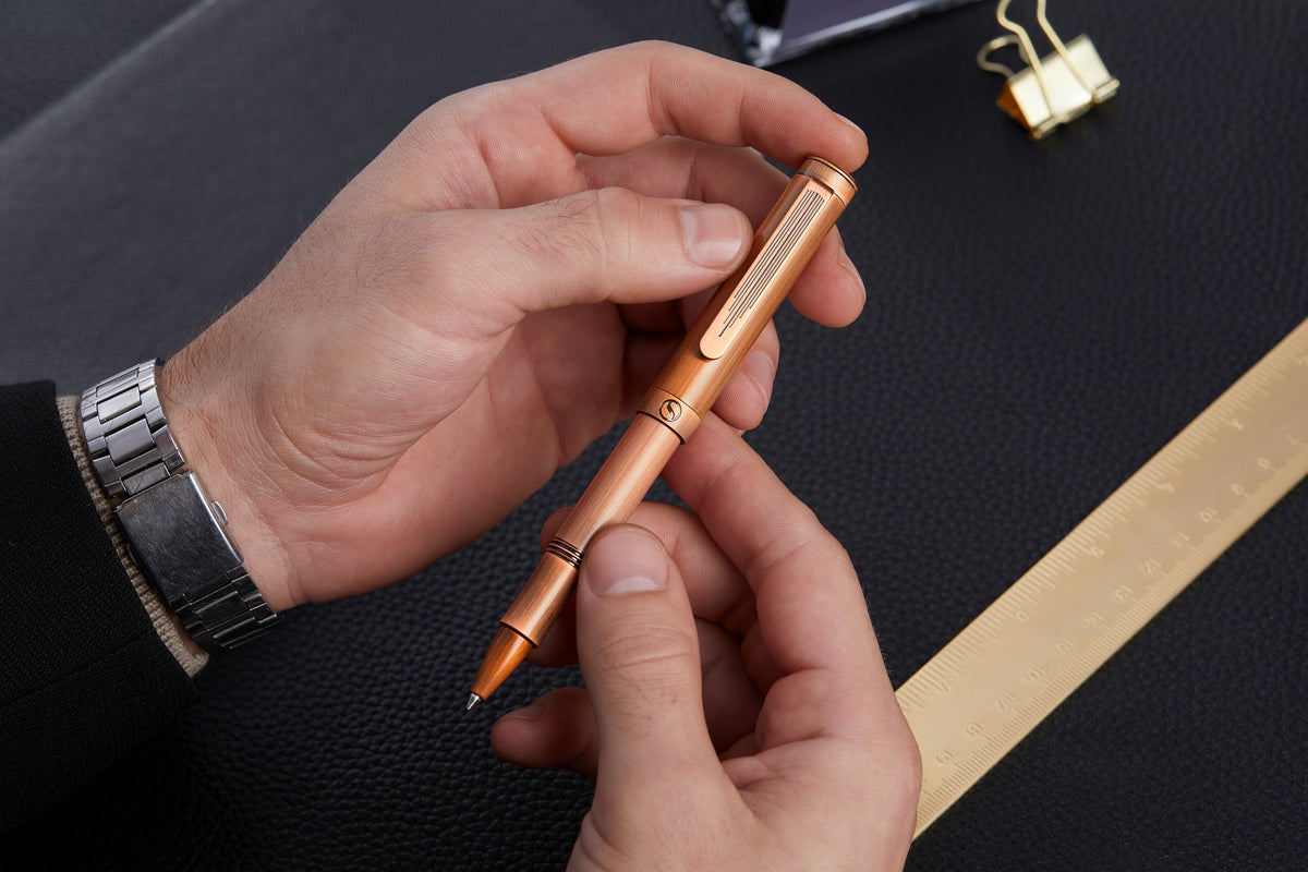 Hand holding a copper pen on a dark surface with a gold ribbon