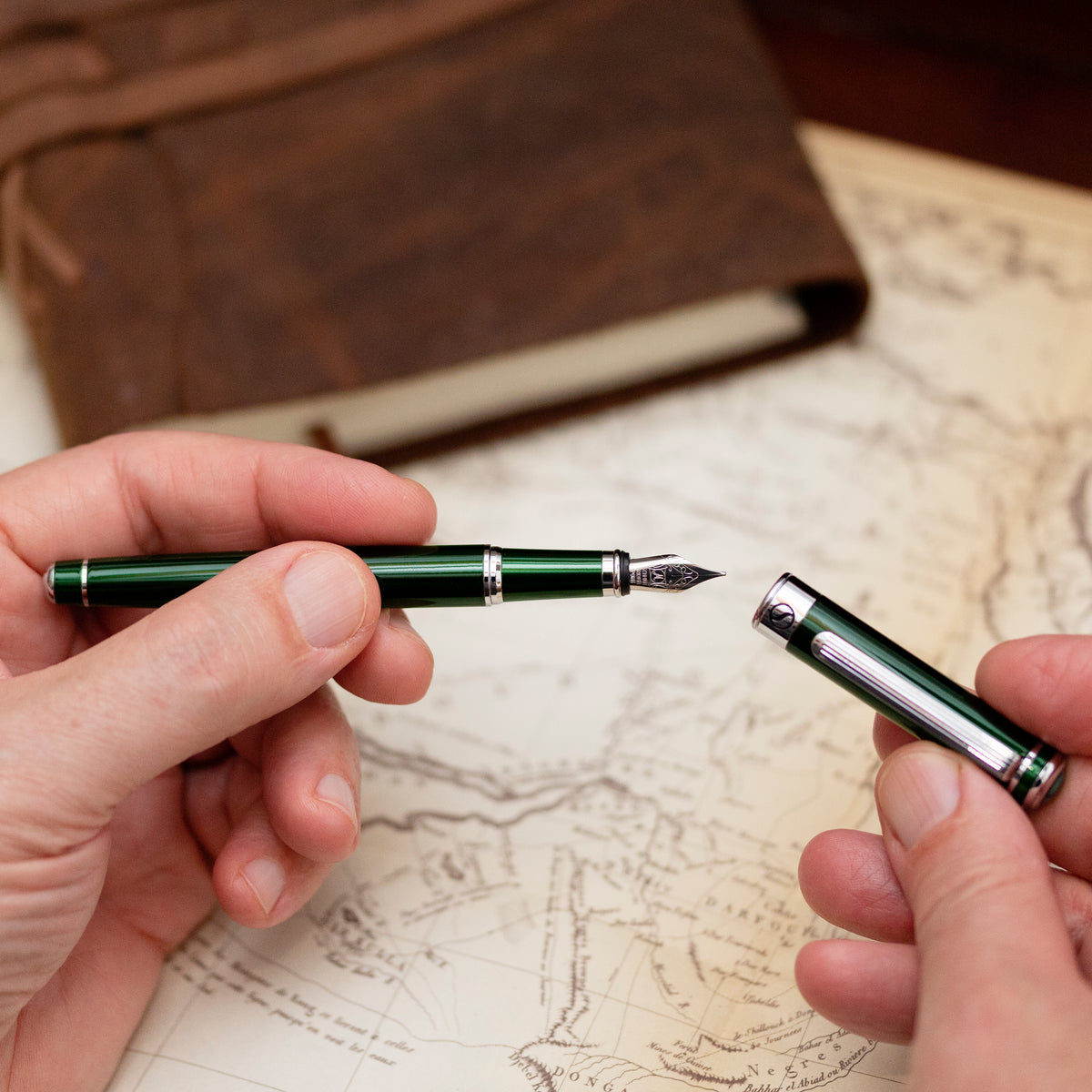 Two hands holding a green fountain pen and its nib against an old map background