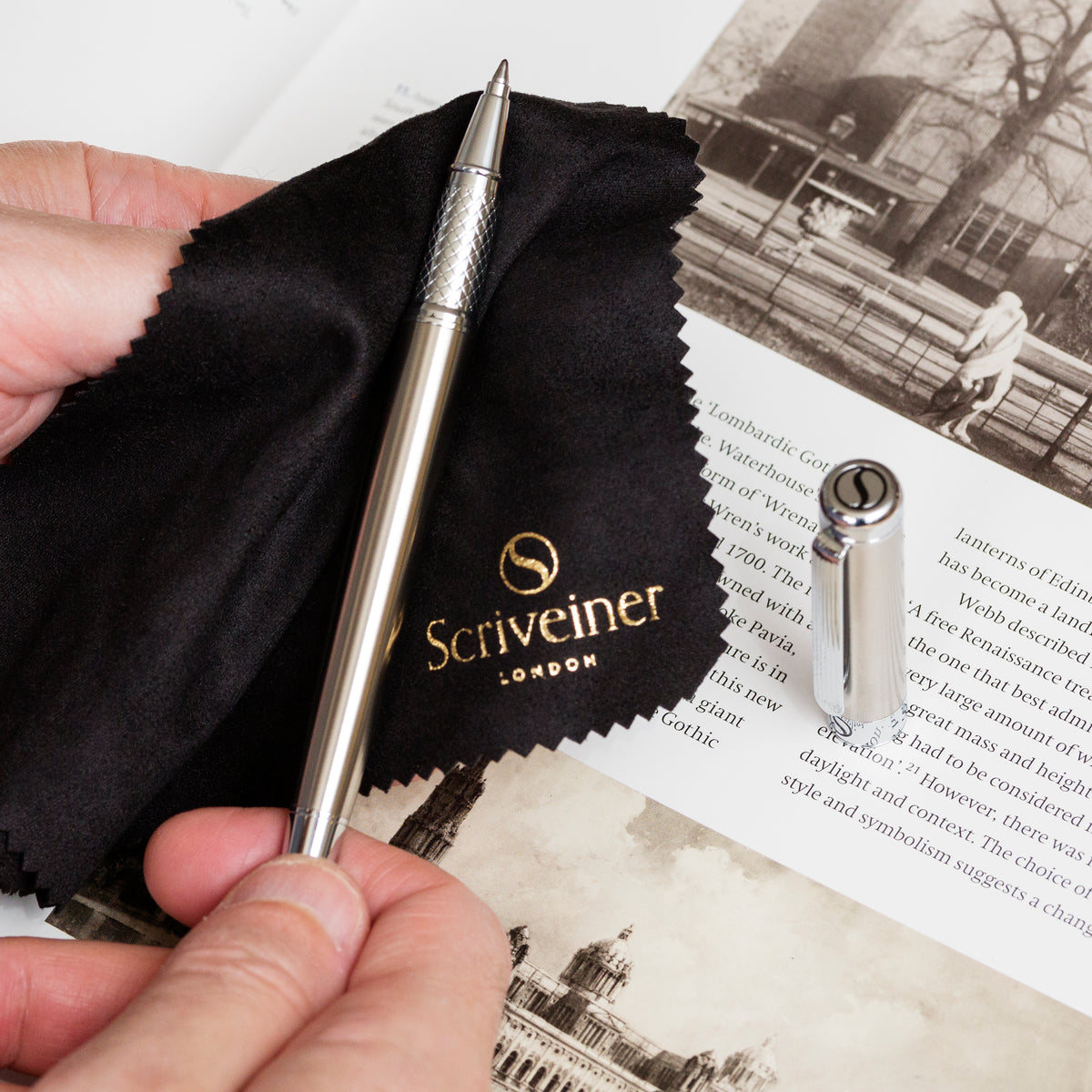 Hand holding a silver pen with 'Scriveiner London' branding on a black cloth and vintage book pages.